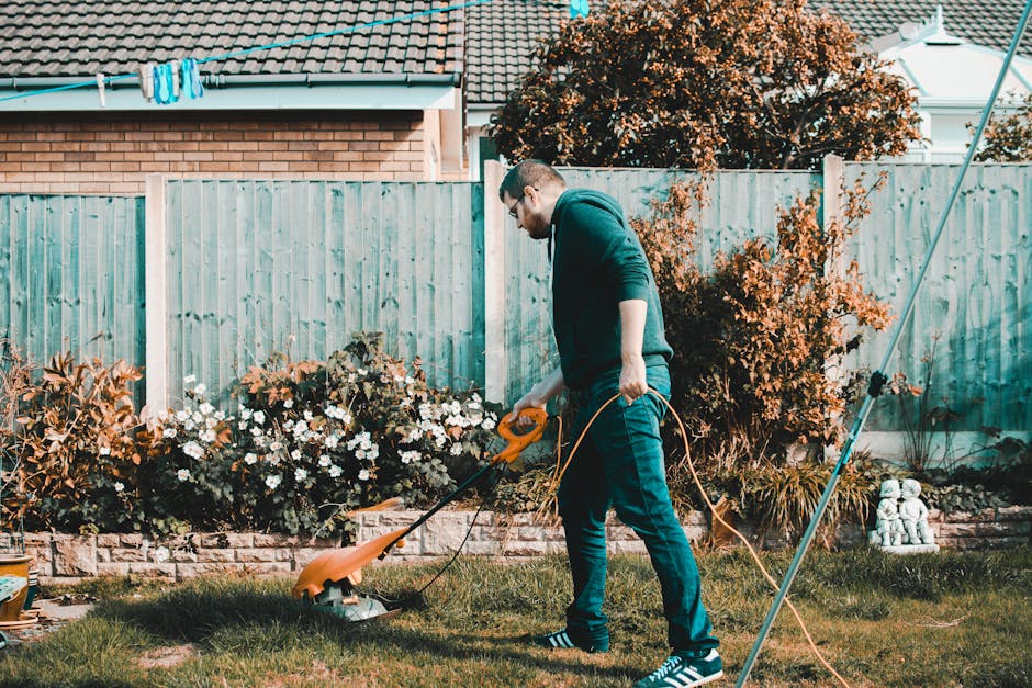 A man wearing a dark hoodie, jeans, and striped sneakers is operating a garden lawnmower on a lush green lawn in a residential garden. The garden features a flower bed with white and orange blossoms, 