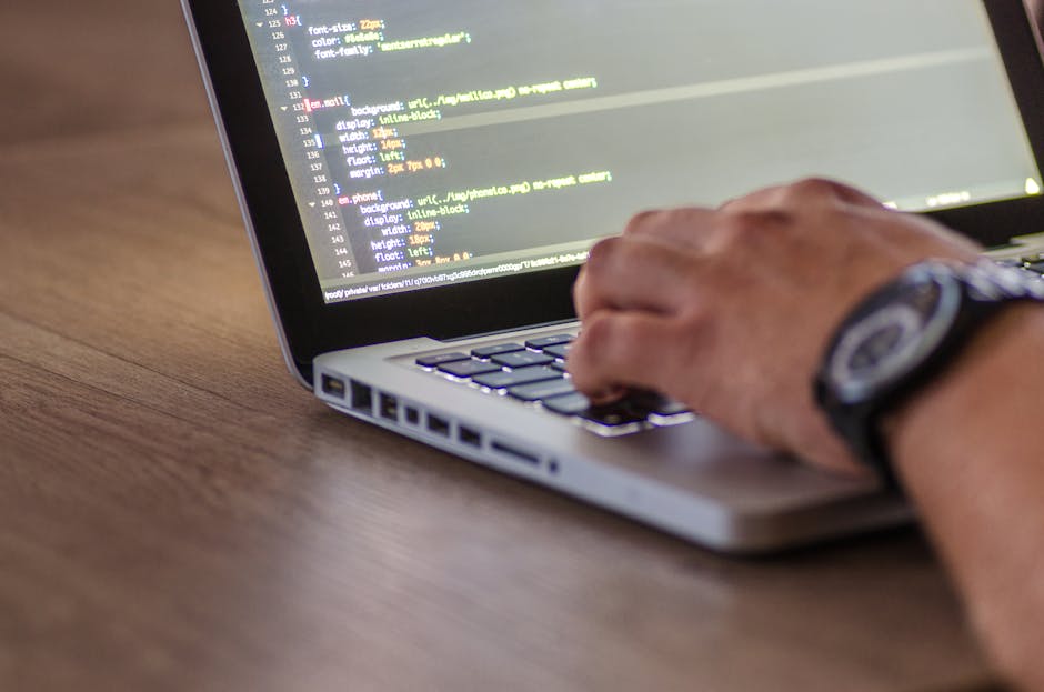 A close-up of a person's hand typing on a silver laptop keyboard placed on a wooden desk. The laptop screen displays lines of colorful code or programming language, with the person's wrist wearing a b