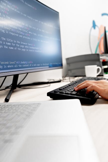 A close-up of a person's hand typing on a silver laptop keyboard placed on a wooden desk. The laptop screen displays lines of colorful code or programming language, with the person's wrist wearing a b